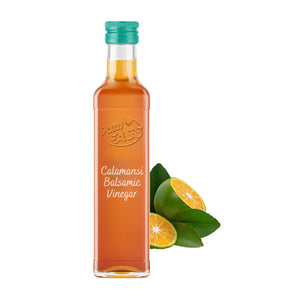 A bottle of Calamansi Balsamic Vinegar with a green cap, accompanied by whole and sliced calamansi fruits with green leaves on a white background.